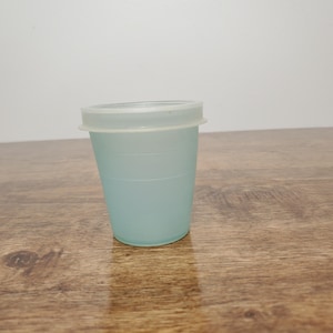 May include: A light blue plastic cup with a white lid. The cup is cylindrical and has horizontal ridges. The lid sits on top of the cup. The cup is on a wooden surface.