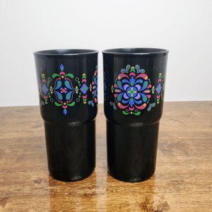 May include: Two black tumblers with colorful floral designs. Each tumbler features a symmetrical pattern with blue, pink, green, and yellow accents. The tumblers are cylindrical and appear to be made of a durable material.