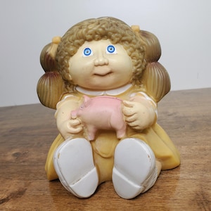 May include: A vintage Cabbage Patch Kids doll figurine in a seated position, holding a pink piggy bank. The doll has blonde curly hair, blue eyes, and is wearing a yellow dress with white shoes. The figurine is made of a hard plastic material.