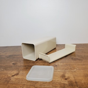 May include: A rectangular, cream-colored plastic storage container with an open end, a matching cream-colored tray insert with raised edges, and a separate, translucent, square-shaped plastic lid resting flat on the surface.
