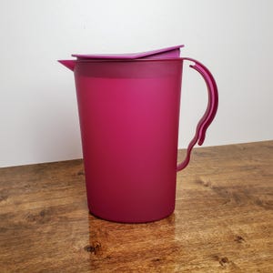 May include: A magenta plastic pitcher with a lid and a handle. The pitcher is on a wooden surface.