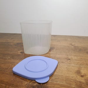 May include: A translucent, square-shaped storage container with vertical ribbed detailing on the sides, accompanied by a separate, flat, light purple lid featuring a circular indentation in the centre and a small tab for easy removal.
