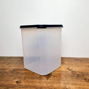 May include: A clear, rectangular food storage container with a black lid. The container is made of translucent plastic and sits on a wooden surface. The lid is flat and fits snugly on top. The container is empty.