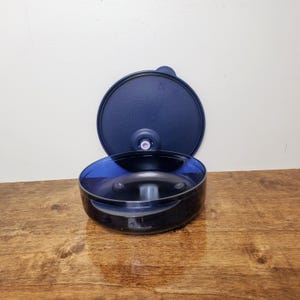 May include: A dark blue plastic container with a matching lid. The container is round with a clear blue interior. The lid is slightly open, revealing the interior. The container sits on a wooden surface.