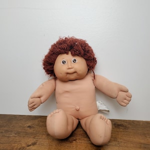 May include: A Cabbage Patch Kids doll with a soft, light-toned fabric body and a round, vinyl head. The doll features short, curly reddish-brown yarn hair, brown eyes, and a small belly button. A white fabric tag is visible on the doll's side.