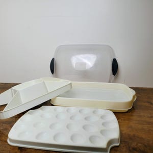 May include: A white plastic food storage container set featuring a rectangular base, a removable divided tray insert, a white egg tray with twenty-four circular indentations, and a clear lid with black side handles for secure closure.