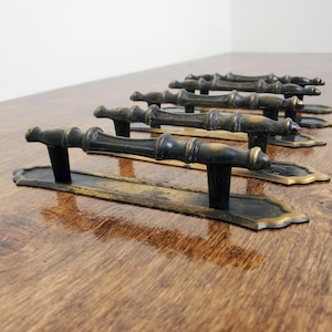 May include: A set of antique bronze door handles. Each handle features a decorative, elongated backplate with a detailed handle. The handles have a dark bronze finish with gold accents, and are arranged on a wooden surface.