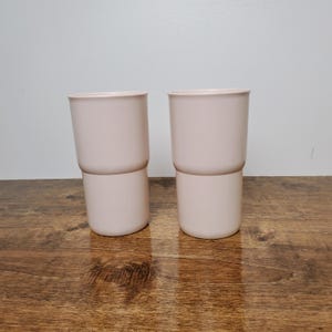 May include: Two pink plastic cups with a double-walled design. The cups are sitting on a wooden surface.