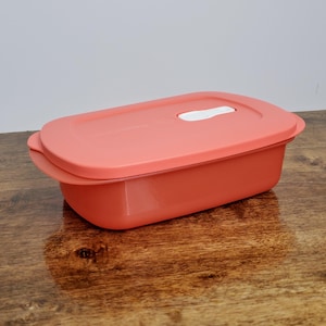 May include: A coral-colored rectangular food storage container with a matching lid. The lid has a white handle. The container is made of plastic and sits on a wooden surface.