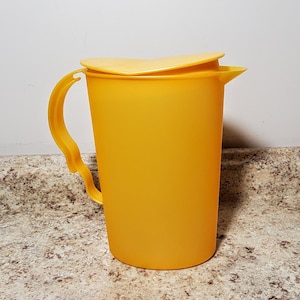 May include: A yellow plastic pitcher with a lid and a handle. The pitcher is made of a smooth, opaque plastic.