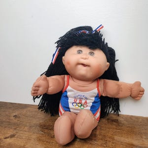 May include: A doll with black yarn hair tied with a red, white, and blue ribbon. The doll has a light skin tone, rosy cheeks, and is wearing a white, blue, and red leotard featuring the text "OlympiKids 1996 USA" and the five Olympic rings.