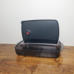 May include: A rectangular, translucent black plastic food storage container with a matching lid. The lid features a circular black vent with a small red center button. The container base is divided into two sections by a central partition.