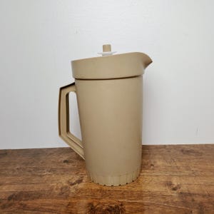 May include: A tan-colored plastic pitcher with a handle and a lid. The pitcher has a spout for pouring and a ridged base. The lid is white and has a small handle. The pitcher is sitting on a wooden surface.