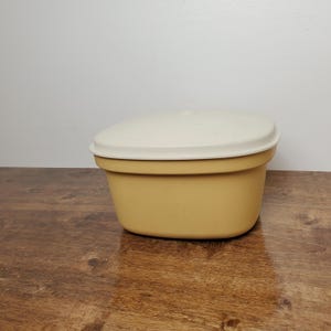 May include: A vintage, mustard-yellow plastic container with a cream-colored lid. The container has a rounded rectangular shape and is sitting on a wooden surface. The lid fits snugly on top, creating a sealed storage space.