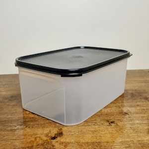 May include: A clear plastic rectangular storage container with a black lid. The container is empty and sitting on a wooden surface.