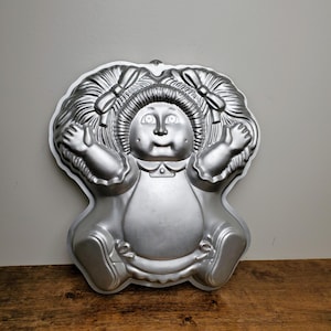May include: A silver metal cake pan shaped like a doll with pigtails. The doll has a round face with large eyes and a wide smile.