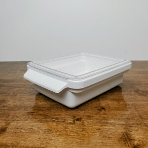 May include: A white rectangular food storage container with a clear lid. The container has a handle on one side and is sitting on a wooden surface. The lid is transparent, allowing a view of the contents inside.