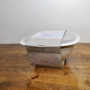 May include: A white Nordic Ware popcorn popper bowl with a translucent lid and a paper product sleeve featuring the text POPCORN POPPER, 12-cup capacity, and an image of popcorn. The bowl has a wide rim and a rounded base for microwave use.