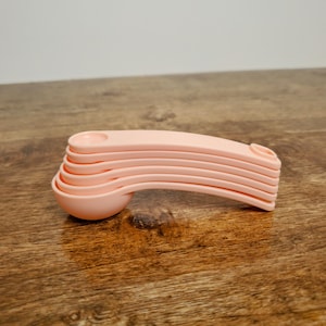 May include: A set of five pink plastic measuring spoons stacked on top of each other. The spoons are all different sizes and have a rounded shape.