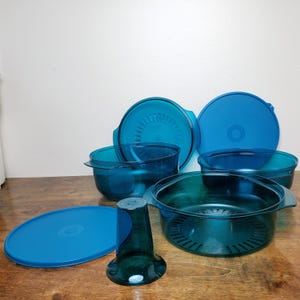 May include: A collection of teal-colored kitchenware, including bowls, lids, and a juicer. The bowls vary in size and shape, with some featuring handles and others having a strainer design. The lids are round and fit the bowls. The juicer is cone-shaped.