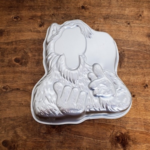 May include: A silver metal cake pan shaped like a furry monster with a large head and two hands. The monster is smiling and has a large, open mouth.