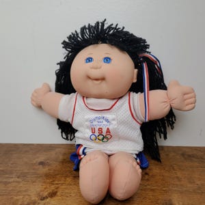 May include: A Cabbage Patch Kids doll with black yarn hair and blue eyes, wearing a white mesh tank top with the text "Olympikids 1996 Official Team Mascot USA" and the Olympic rings logo, paired with blue and white athletic shorts with red trim.