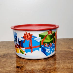 May include: A white plastic container with a red lid. The container has a Christmas theme with a design of presents wrapped in red, blue, and green paper with bows.