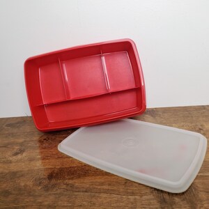 May include: A red plastic food storage container with three compartments, and a separate, translucent lid. The container is rectangular with rounded corners. The lid is also rectangular with rounded corners and a circular design in the center.