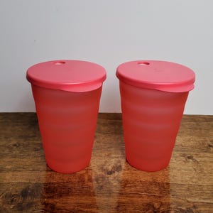 May include: Two red plastic tumblers with matching pink lids. Each tumbler has a ridged design and a small hole in the center of the lid. The tumblers are sitting on a wooden surface.