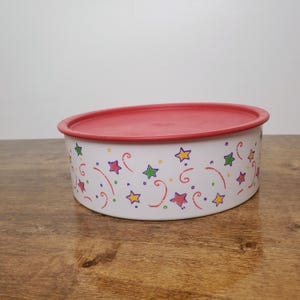 May include: A round white storage container with a red lid. The exterior of the container features a repeating pattern of colorful stars in yellow, purple, green, and red, interspersed with small red and orange swirling line accents.