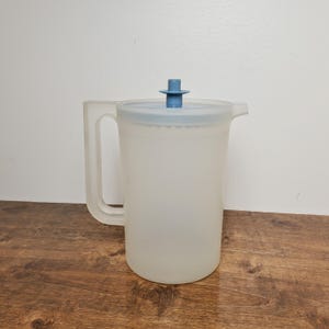 May include: A translucent plastic pitcher with a light blue lid and handle. The pitcher has a capacity of approximately 64 ounces. The lid features a blue, cross-shaped handle. The pitcher is sitting on a wooden surface.