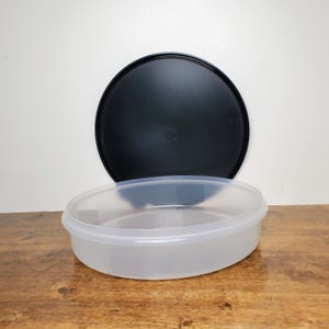May include: A clear, oval-shaped plastic container with a black lid. The container is translucent, allowing visibility of the contents. The lid is round and sits above the container. The items are on a wooden surface.