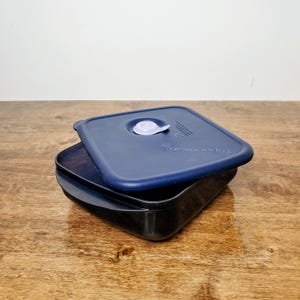 May include: A dark blue Tupperware container with a matching lid. The lid has a white valve and is slightly ajar, revealing the black interior of the container. The container sits on a wooden surface.