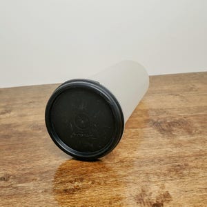 May include: A white plastic container with a black lid. The lid has a small circle in the center.