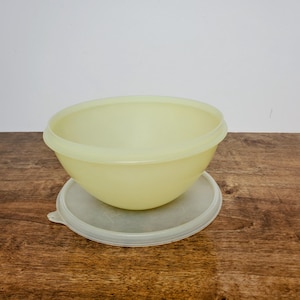 May include: A pale yellow plastic mixing bowl with a matching lid. The bowl is round with a slightly flared rim and sits atop a clear, round lid. The bowl and lid are on a wooden surface.