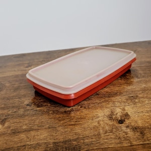 May include: A red plastic rectangular container with a clear lid. The container is on a wooden surface.
