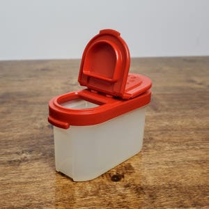 Pu&ograve; includere: Un contenitore per spezie in plastica rossa e trasparente con un coperchio incernierato. Il coperchio &egrave; aperto e il contenitore &egrave; su una superficie di legno.