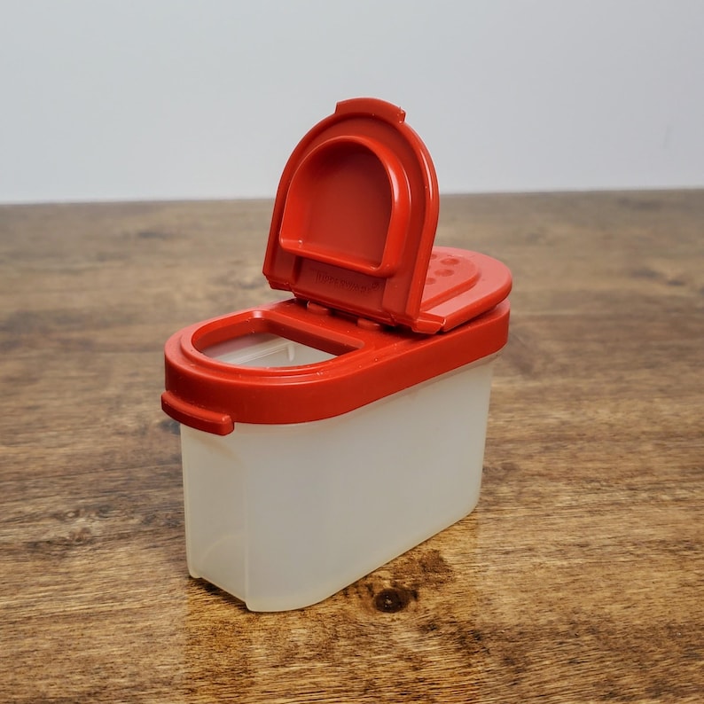 Pu&ograve; includere: Un contenitore per spezie in plastica rossa e trasparente con un coperchio incernierato. Il coperchio &egrave; aperto e il contenitore &egrave; su una superficie di legno.