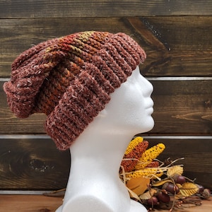 May include: A brown, orange, and yellow knitted beanie hat with a slouchy fit. The hat is displayed on a white mannequin head.