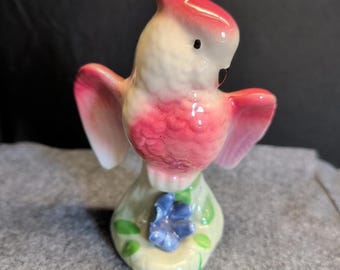 May include: A ceramic figurine of a bird with a pink crest, wings, and body, and a white face. The bird is perched on a light green base with a blue flower and green leaves. The figurine is a decorative item.