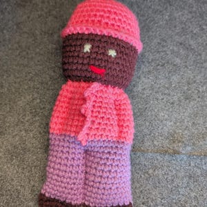 May include: A handmade crochet doll with a pink hat, jacket, and purple pants. The doll's face is brown with white eyes and a red mouth. The doll is made of yarn and has a textured appearance.