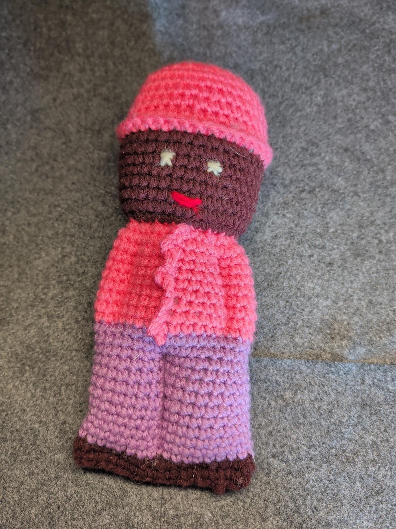 May include: A handmade crochet doll with a pink hat, jacket, and purple pants. The doll's face is brown with white eyes and a red mouth. The doll is made of yarn and has a textured appearance.