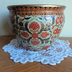 Puede incluir: Una maceta de cerámica decorativa con un diseño floral en rojo, negro y dorado. La maceta se encuentra sobre un tapete de encaje blanco. El borde de la maceta tiene una banda dorada.