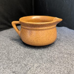 May include: A small, speckled, tan-colored ceramic creamer with a handle and a spout. The creamer has a rounded body and a slightly flared rim. The handle is small and curved, and the spout is pointed. The creamer is likely vintage.