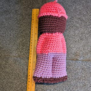 May include: A handmade crochet doll in the shape of a cupcake. The doll features a pink top, a dark brown middle section, a pink body, and purple legs. A wooden ruler is placed next to the doll.