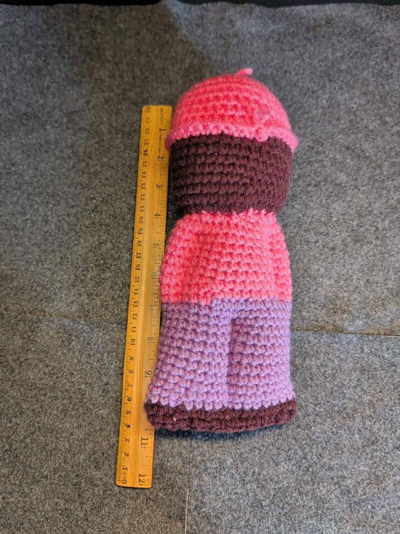 May include: A handmade crochet doll in the shape of a cupcake. The doll features a pink top, a dark brown middle section, a pink body, and purple legs. A wooden ruler is placed next to the doll.