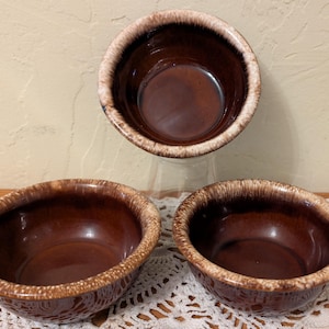 May include: Three round ceramic bowls featuring a deep, glossy brown interior and exterior glaze. Each bowl has a distinct, textured rim with a mottled cream and light brown finish that contrasts with the smooth, dark brown body of the vessel.