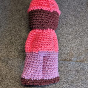 May include: A handmade crochet doll in shades of pink, purple, and brown. The doll features a rounded head, a dark brown face, a pink body, and purple legs. The doll is made with a tight crochet stitch.
