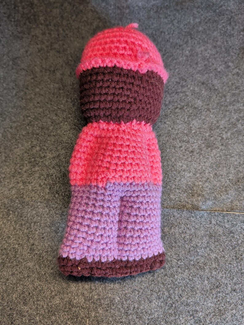 May include: A handmade crochet doll in shades of pink, purple, and brown. The doll features a rounded head, a dark brown face, a pink body, and purple legs. The doll is made with a tight crochet stitch.
