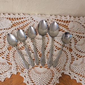 Puede incluir: Un juego de seis cucharas de metal plateado dispuestas en fila. Cada cuchara presenta un cuenco liso y pulido, y un mango con un patrón decorativo en relieve en la base, que incluye una letra R estilizada grabada en las cucharas más grandes.